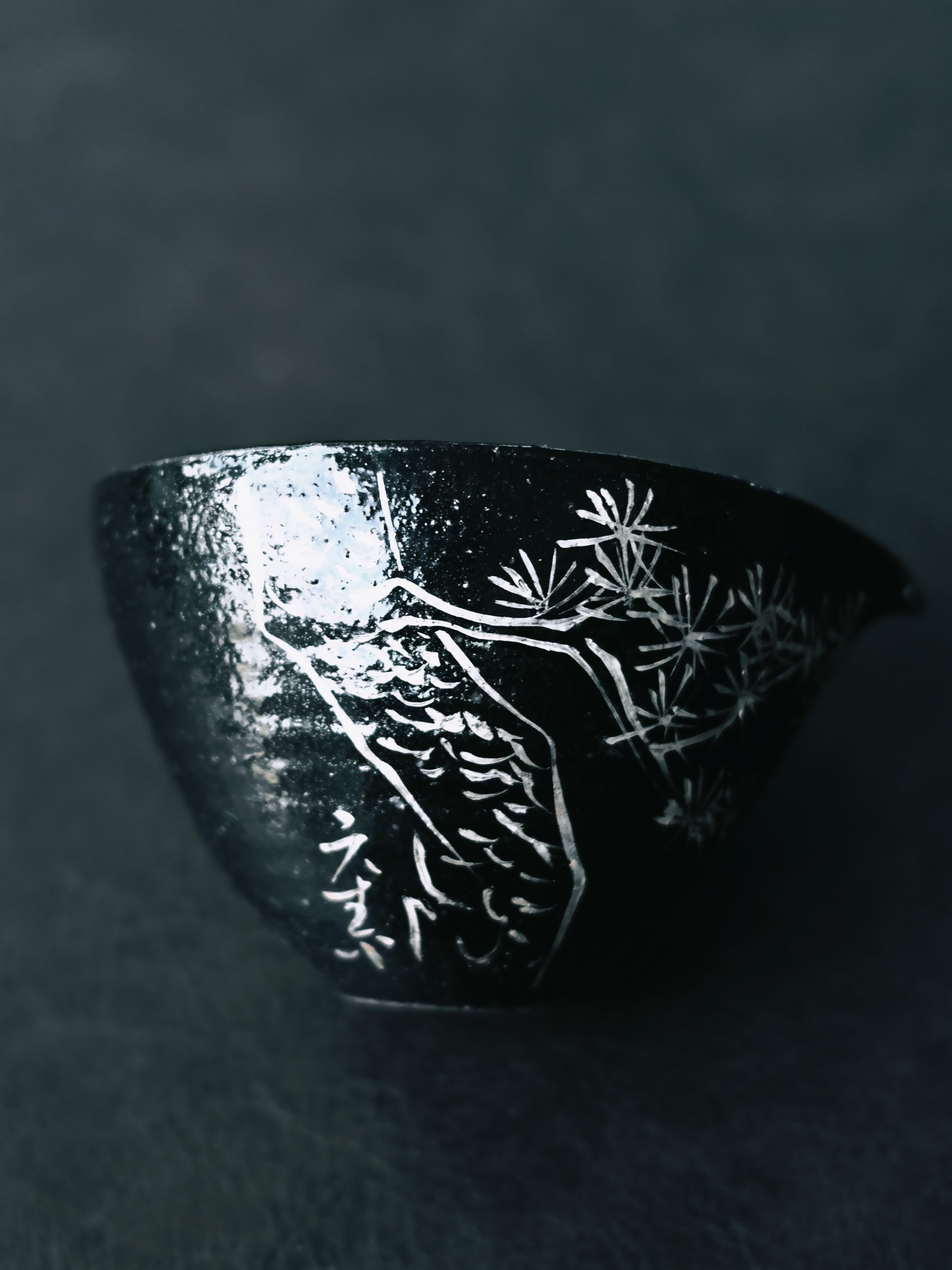 Artisanal raku-inspired set with glossy black glaze, hand-applied silver poem fragments and pine silhouettes, deliberately fractured and repaired in kintsugi spirit.