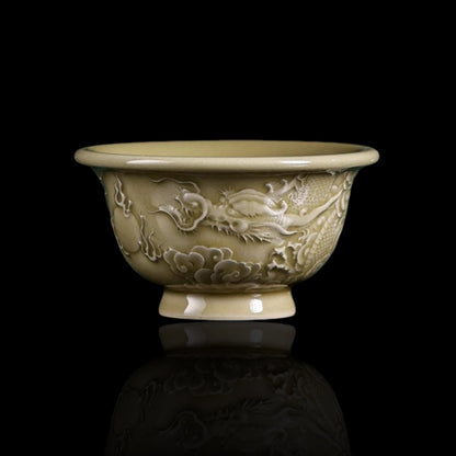 A close-up view of the Imperial Longquan Celadon tea cup featuring intricate hand-carved dragon relief against a dark green gradient background.