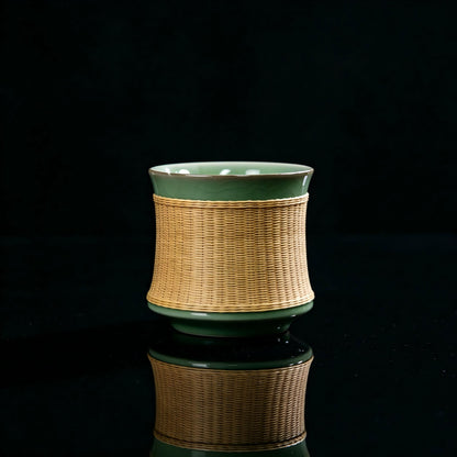 Main product shot of Longquan Ge‑Yao celadon teacup with hand‑woven bamboo sleeve, plum‑green crackle glaze, black background with mirror reflection