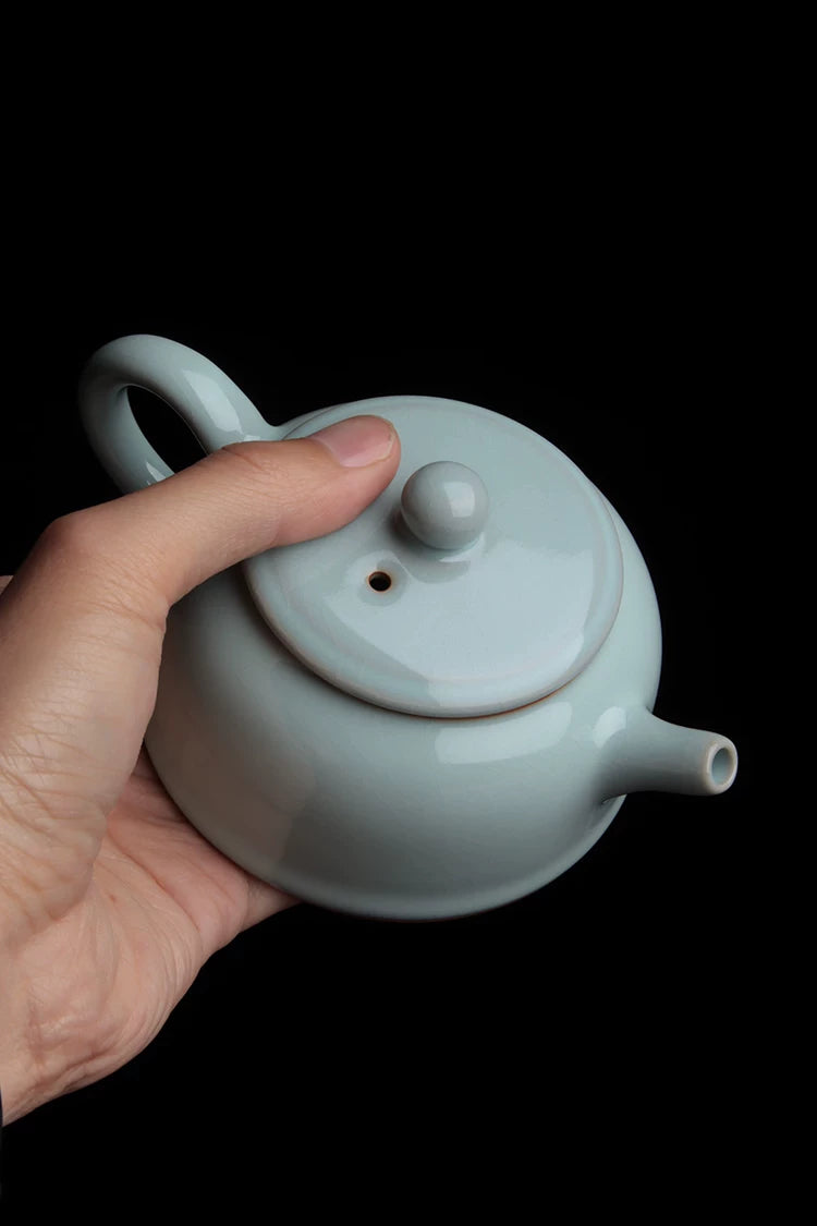 A hand holding the sky-cyan Ru Kiln teapot to demonstrate comfortable grip and proportion.