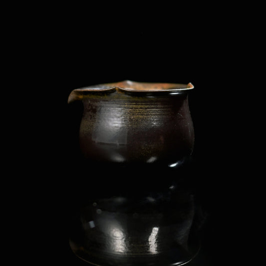 Unique piece wood-fired black gold mud fairness cup on a rustic wood slice against a green backdrop.